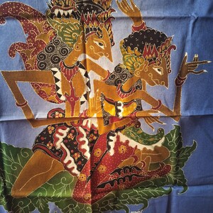 Peut inclure: Un tissu bleu présentant une impression batik détaillée de deux personnages stylisés portant des coiffes traditionnelles et des vêtements ornés. Les personnages sont représentés dans une pose dynamique sur fond bleu, avec le mot SANDI en blanc.