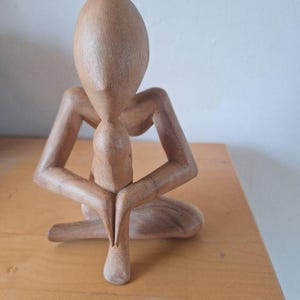 Op de afbeelding: Een lichtbruin houten beeldhouwwerk van een gestileerde menselijke figuur in een zittende, introspectieve houding. De figuur heeft een ovaalvormig hoofd, hoekige schouders en armen die op de knieën rusten, met een gladde, gepolijste houtnerf.