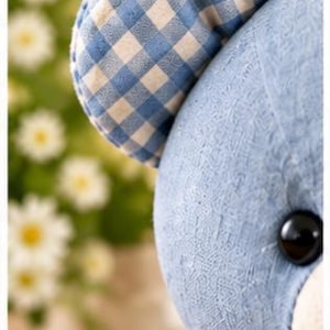 May include: A plush toy bear featuring a blue textured fabric head and a rounded ear with a blue and white gingham check pattern. The bear has a shiny black button eye and a light-coloured muzzle. The text 6. Ear Detail is visible.