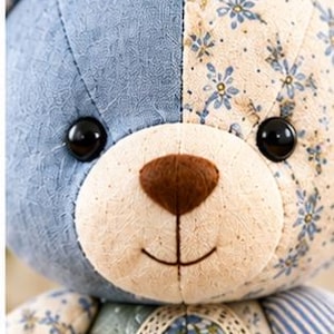 May include: A stuffed bear toy featuring a patchwork design with blue and cream fabric panels. The bear has black button eyes, a brown felt nose, and a stitched mouth. The ears are blue and white checkered, and the chest has lace trim.