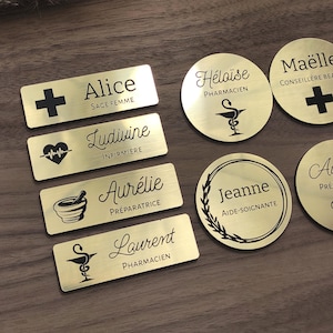 Op de afbeelding: Een verzameling rechthoekige en ronde naamplaatjes van goudkleurig metaal met gegraveerde zwarte tekst en medische symbolen. De plaatjes tonen namen zoals Alice, Ludivine, Aurélie, Laurent, Héloïse, Maëlle en Jeanne, samen met hun professionele titels.