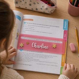 Peut inclure: Un guide de lecture rectangulaire en plastique rose translucide portant le prénom Charline écrit en lettres cursives blanches, orné de petites fleurs dorées, posé horizontalement sur une page de manuel scolaire en français.