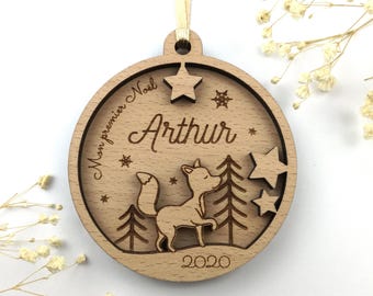 Puede incluir: Un adorno navideño circular de madera clara con un grabado láser de un zorro, pinos, estrellas y copos de nieve. El texto dice "Mon premier Noël", "Arthur" y "2020". Una cinta de color claro está sujeta al lazo superior.