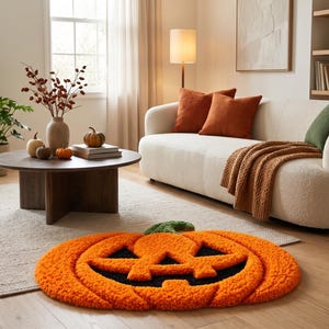 Op de afbeelding: Een rond, getextureerd vloerkleed in de vorm van een Halloween-pompoen. Het kleed heeft een feloranje buitenkant met een zwart uitgesneden gezichtspatroon en een kleine groene steel aan de bovenkant. Het oppervlak heeft een getufte structuur.