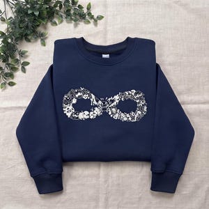 Puede incluir: Una sudadera azul marino de manga larga con un gran estampado gráfico blanco en el pecho. El diseño tiene forma de símbolo de infinito y está compuesto por numerosas ilustraciones pequeñas y detalladas de personajes y garabatos.