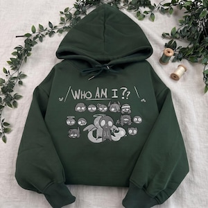 Puede incluir: Una sudadera con capucha de color verde oscuro que presenta un estampado gráfico con el texto "WHO AM I??" sobre una colección de varias ilustraciones pequeñas y estilizadas de personajes en tonos gris, blanco y negro, incluida una figura alada central.