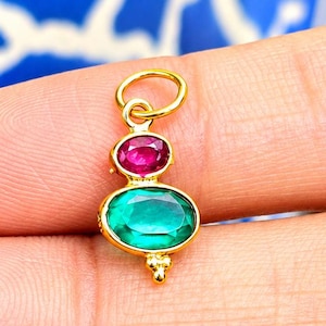 Pode incluir: Um pingente de ouro com uma pequena pedra preciosa rosa de formato oval posicionada acima de uma pedra verde oval maior. O pingente inclui um anel de salto circular de ouro no topo e uma pequena conta decorativa de ouro na base do engaste.