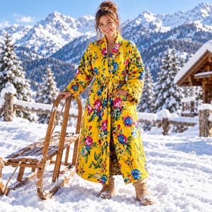 Puede incluir: Una bata larga acolchada de tela amarilla vibrante con un colorido estampado floral de flores rosas, azules y verdes. La prenda cuenta con mangas largas, cuello tipo chal y un cinturón a juego en la cintura, llegando hasta los tobillos.