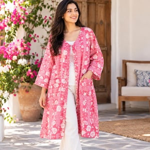 Könnte beinhalten: Ein langärmeliger, vorne offener Mantel in leuchtendem Pink mit einem durchgehenden weißen Blumenmuster. Das Kleidungsstück verfügt über zwei aufgesetzte Vordertaschen, reicht bis zum Knie und wird über einem weißen Oberteil und einer Hose getragen.