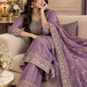Può includere: Un completo color lavanda composto da una tunica a maniche lunghe, pantaloni a gamba larga e una dupatta abbinata. Il tessuto presenta un motivo floreale tono su tono con intricati ricami dorati e perline lungo i bordi e lo scollo.