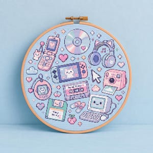 以下が含まれることがあります： 水色の布にクロスステッチが施された円形の木製刺繍枠。パステルカラーのレトロな電子機器がデザインされており、折りたたみ式携帯電話、CD、猫耳ヘッドフォン、携帯ゲーム機、カセットテープ、インスタントカメラ、フロッピーディスク、コンピューターなどが描かれています。