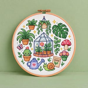 May include: A circular wooden embroidery hoop containing a cross-stitch pattern of various plants, a greenhouse, a snail, a watering can, and a seed packet labelled SEEDS. The design features potted plants, a hanging plant, a mushroom, and a butterfly.