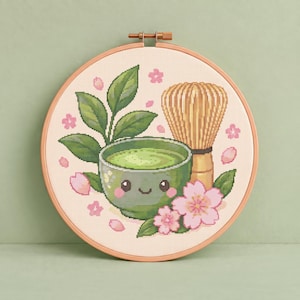 Könnte beinhalten: Ein Kreuzstichmuster in einem hölzernen Stickrahmen mit einer grünen Matcha-Schale mit lächelndem Gesicht, einem Bambusbesen, grünen Teeblättern und rosa Kirschblütenblättern auf einem hellen, cremefarbenen Stoffhintergrund.