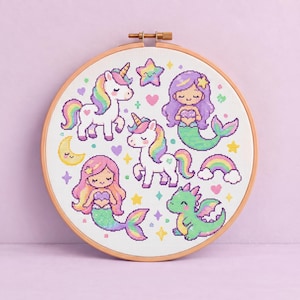 May include: A circular wooden embroidery hoop containing a white fabric piece with cross-stitch designs of two white unicorns with rainbow manes, two mermaids with colorful tails, a green dragon, a yellow crescent moon, and a rainbow.