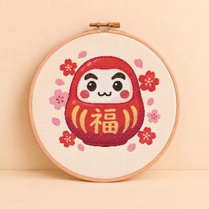 May include: A circular wooden embroidery hoop containing a cross-stitch pattern of a red Daruma doll with a white face, black eyes, and a gold Japanese character for luck. The doll is surrounded by small red and pink cherry blossom flowers.