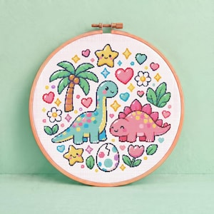 May include: A circular wooden embroidery hoop containing a cross-stitch pattern of a blue long-necked dinosaur and a pink stegosaurus. The design includes a palm tree, a cracked egg, flowers, hearts, and yellow stars on a white fabric background.