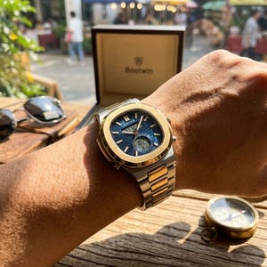 Op de afbeelding: Een tweekleurig polshorloge met een schakelarmband van zilver- en goudkleurig roestvrij staal en een blauwe wijzerplaat met horizontale strepen. Het horloge heeft een goudkleurige achthoekige lunette, analoge wijzers en een digitaal display.