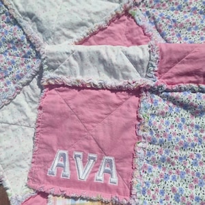 May include: A patchwork rag quilt featuring alternating squares of solid pink fabric, white fabric with tiny pink floral patterns, and a multi-coloured floral print with blue and pink flowers. One pink square features the name AVA in white embroidery.