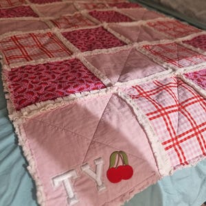 May include: A handmade rag quilt featuring a patchwork of pink, white, and red fabrics with various patterns including plaid and small prints. The quilt has frayed seams and includes the embroidered white letters TY and a red cherry applique.
