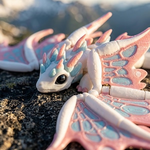 May include: A small, articulated dragon figurine featuring a white body with pastel pink and light blue accents on its wings, head, and back. The creature has large, dark, glossy eyes and a textured surface, with wings that display a delicate, vein-like pattern in pink and blue.