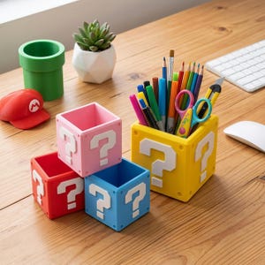 以下が含まれることがあります： 黄色、ピンク、赤、青のクエスチョンマークブロックの形をした正方形のデスクオーガナイザーセット。黄色いブロックには、色鉛筆、ペン、ハサミ、カッターナイフが入っています。緑色のパイプ型のカップと赤い帽子が近くに置かれています。