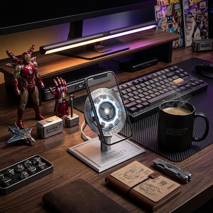 May include: A wireless phone charging stand featuring an illuminated arc reactor design with the text PROOF THAT TONY STARK HAS A HEART. The base is labelled INFINITY SAGA. The charger is positioned on a desk with a black mug, a keyboard, and various figurines.