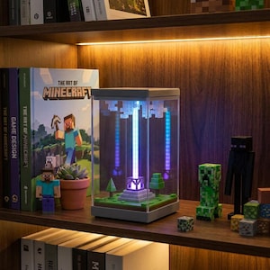 Może przedstawiać: Prostokątna lampka Minecraft w kształcie latarni z przezroczystą obudową z pleksi, szarą podstawą i centralnym pionowym strumieniem światła emitującym jasnoniebieski blask. Wewnątrz znajduje się miniaturowy, klockowy krajobraz z zielonymi drzewami.