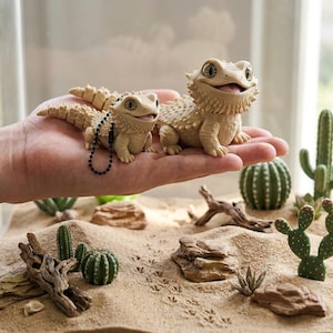 May include: Two small, beige, textured lizard figurines with large, dark, expressive eyes and open mouths. One figurine features a black beaded chain attached to its side, while both display detailed scales and a spiky appearance on their bodies.