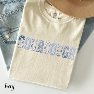 Peut inclure: Un t-shirt à manches courtes couleur ivoire portant le mot SOURDOUGH en grandes lettres style bloc. Chaque lettre est remplie d'un tissu différent à motifs bleus et blancs, incluant des designs floraux, vichy et à pois sur le coton.