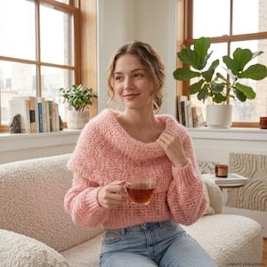 May include: A soft, light pink, chunky knit sweater with a wide, folded-over collar and long sleeves, paired with blue denim jeans. The sweater features a textured, open-weave pattern and a cropped fit, worn while holding a clear glass mug of tea.