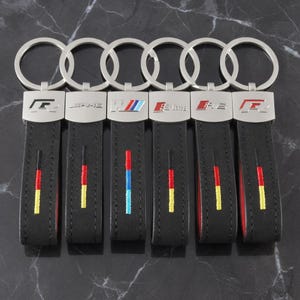 Puede incluir: Seis llaveros de cuero negro con herrajes de metal plateado y anillas circulares. Cada llavero presenta un logotipo automotriz único, incluyendo R, AMG, M, S-line y RS. Las correas de cuero están bordadas con rayas verticales de varios colores.