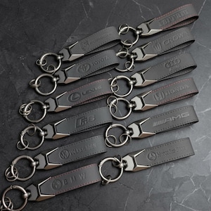Peut inclure: Une collection de porte-clés en cuir gris foncé arborant divers logos de marques automobiles dont Ferrari, Porsche, Crown, Toyota, Audi, Lexus, Honda, RS, AMG, Volkswagen, Mercedes-Benz et BMW, chacun avec un fermoir et un anneau métalliques.