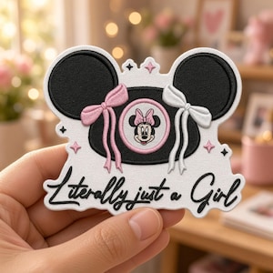 May include: An embroidered patch featuring a black Minnie Mouse silhouette with a pink bow on the left and a white bow on the right. A small circular portrait of Minnie Mouse is in the center. The text "Literally just a Girl" is written below.