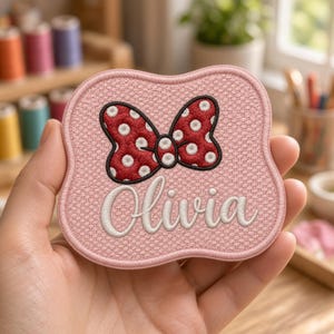 May include: A pink embroidered patch with a scalloped edge featuring a red bow with white polka dots and the name Olivia stitched in white cursive script. The patch has a textured fabric surface and a black border around the bow design.
