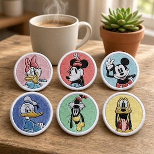 May include: Six circular embroidered patches featuring Disney characters: Daisy Duck on pink, Minnie Mouse on coral, Mickey Mouse on light blue, Donald Duck on blue, Goofy on light green, and Pluto on yellow, all with white borders.