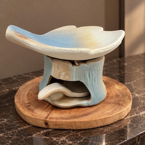May include: A ceramic incense burner featuring a wide, leaf-shaped top dish with a light blue and cream gradient finish, supported by a textured, tree-stump-shaped base in a matching pale blue hue with a small curved ceramic spoon inside.
