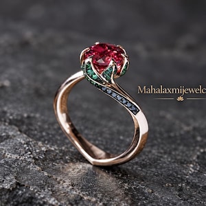 Puede incluir: Un anillo de oro rosa con una gran piedra roja de corte redondo en el centro, rodeada por un engaste floral con pequeñas piedras verdes y negras a lo largo de la banda. El texto Mahalaxmijewelcrafts es visible a la derecha del anillo.