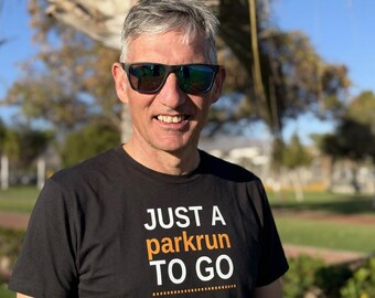May include: A black short-sleeved t-shirt featuring the white and orange text "JUST A parkrun TO GO" printed on the front. The shirt has a crew neckline and a standard fit, made of a solid black fabric with clear, bold lettering across the chest area.