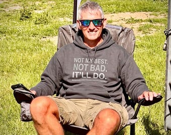 May include: A dark gray hooded sweatshirt featuring the white text "NOT MY BEST. NOT BAD. IT'LL DO." printed on the chest. The garment includes a front kangaroo pocket, long sleeves, and a drawstring hood, worn with khaki-colored shorts.