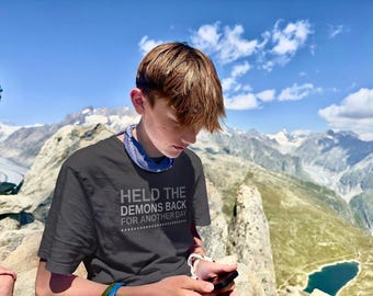 May include: A dark gray short-sleeved t-shirt featuring the white text "HELD THE DEMONS BACK FOR ANOTHER DAY" printed on the chest, accompanied by a blue patterned neck gaiter worn around the neck, set against a backdrop of mountain peaks.