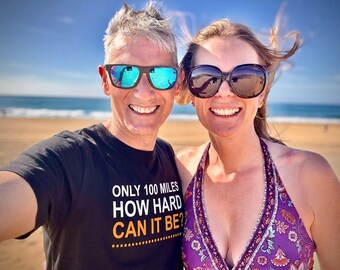 May include: A black t-shirt with the white and orange text ONLY 100 MILES HOW HARD CAN IT BE? and a purple patterned halter top. The t-shirt features a dashed line graphic below the text. Both items are worn by individuals wearing sunglasses.