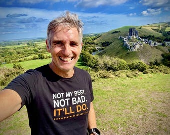 May include: A black short-sleeved t-shirt featuring the white and orange text "NOT MY BEST. NOT BAD. IT'LL DO." printed on the chest. The shirt has a crew neckline and a standard fit, showcasing clear lettering against the dark fabric background.