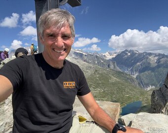May include: A black short-sleeved t-shirt featuring a rectangular orange logo on the left chest with white icons of a runner and a camera, accompanied by the white text "Film My Run" printed directly below the icons on the shirt fabric.