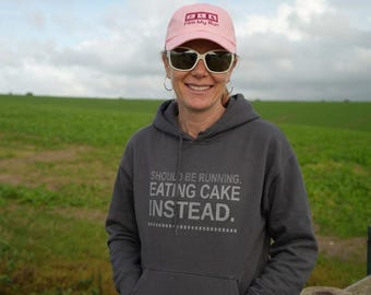 May include: A charcoal gray hooded sweatshirt featuring the text SHOULD BE RUNNING, EATING CAKE INSTEAD. in light gray lettering, paired with a light pink baseball cap with the text Film My Run and white-rimmed sunglasses with dark lenses.