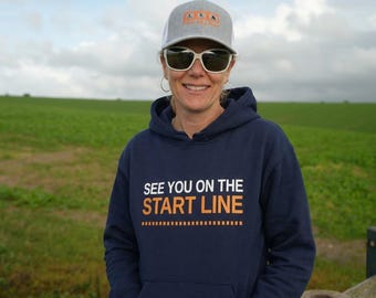 May include: A navy blue hooded sweatshirt featuring the white and orange text "SEE YOU ON THE START LINE" with a dashed line graphic, paired with a grey and white baseball cap with an orange logo and white-framed sunglasses.