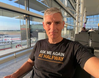 May include: A black short-sleeved t-shirt featuring the white and orange text "ASK ME AGAIN AT HALFWAY" printed on the chest, with a dashed line graphic underneath the text. The shirt has a crew neckline and a standard fit.