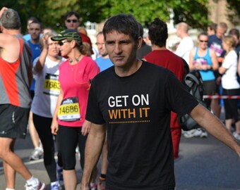 May include: A black short-sleeved t-shirt featuring the white and orange text "GET ON WITH IT" printed on the chest. The shirt has a crew neckline and a standard fit, worn by a runner in a race with other participants visible in the background.