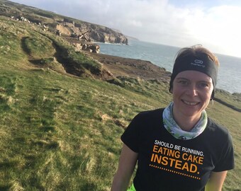 May include: A black short-sleeved t-shirt featuring the white and orange text SHOULD BE RUNNING. EATING CAKE INSTEAD. The shirt is worn with a black headband with white lettering and a light-colored patterned neck gaiter.