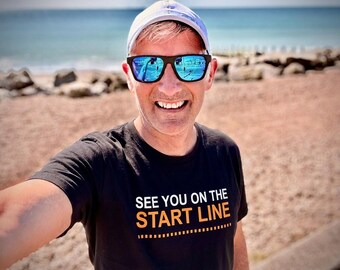May include: A black short-sleeved t-shirt featuring the white and orange text "SEE YOU ON THE START LINE" with a dashed line graphic underneath. The shirt is worn with a white baseball cap and blue-tinted sunglasses with black frames.