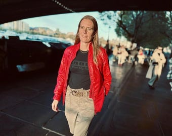 May include: A bright red jacket worn over a black t-shirt with the text Film My Run, paired with tan trousers featuring a wide belt with a decorative buckle. The jacket has a smooth texture and long sleeves, while the trousers have a straight fit.
